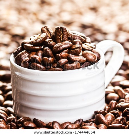 White Cup full of roasted coffee beans on heap of coffee beans background. Coffee background texture with copy space for text, close up.