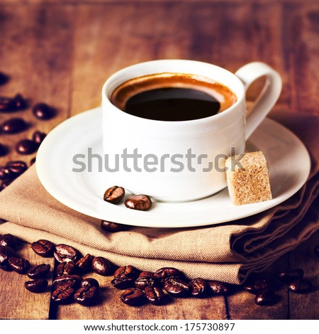 Cup of coffee with coffee beans on wooden table on brown background closeup. Cup of espresso and white saucer on brown napkin with cane sugar.