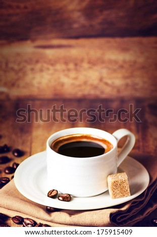Cup of coffee with coffee beans on a beautiful wooden  brown background in rustic style with copyspace for text, closeup. Cup of espresso and white saucer on brown napkin with cane sugar.