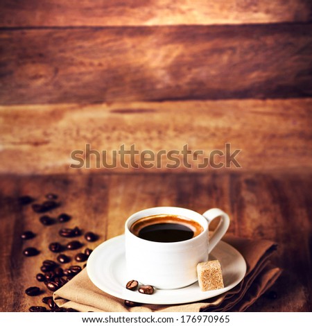 Cup of coffee with coffee beans on a beautiful wooden  brown background in rustic style with copyspace for text, closeup. Cup of espresso and white saucer on brown napkin with cane sugar.