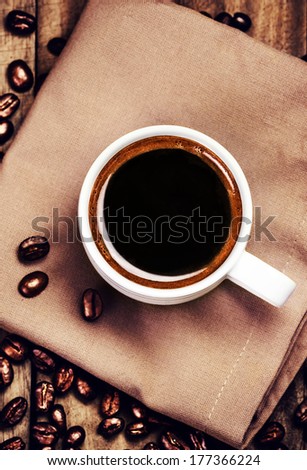 Cup of coffee with coffee beans on  brown napkin on wooden background. White Cup of espresso, top view.