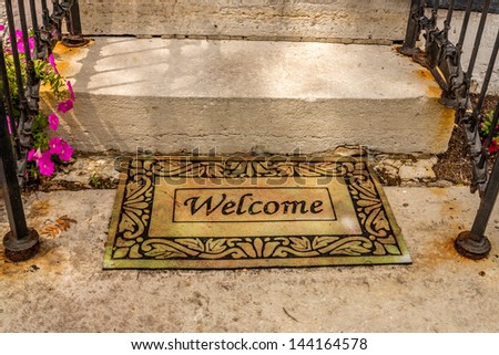 Welcome mat sitting at the bottom of a set of stairs with black hand rails and flowers