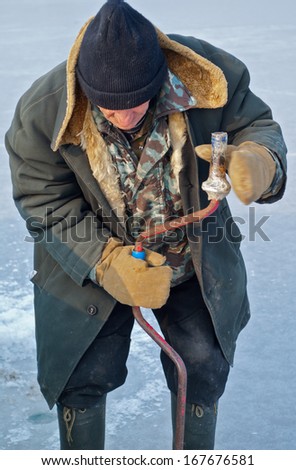 Old man drills ice on winter fishing.