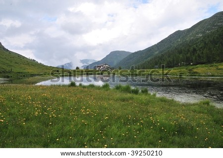 Cloudy landscape with alpine lake (tarn), dandelion (Taraxacum officinale) and a typical building on background