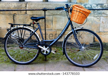 A blue bike leaning against a railing and wall. The bike has a wicker basket attached to the handlebars.