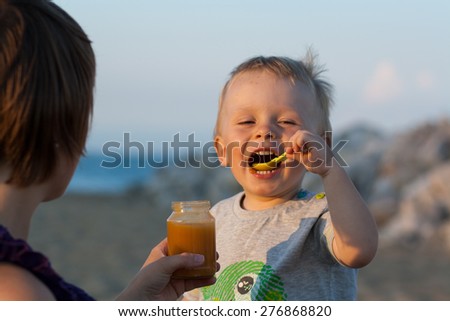 baby is feeding baby food, with green plastic spoon, by the sea, on sand beach, beach with rocks. Mother is feeding a baby. Healthy food.