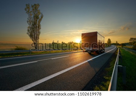 Red small truck driving on asphalt road around farm fields in rural landscape at sunset
