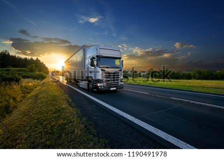 White trucks arriving on the asphalt road in rural landscape in the rays of the sunset