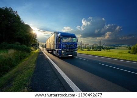 Blue truck arriving on the asphalt road in rural landscape in the rays of the sunset