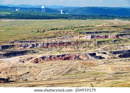 Surface mine with red minerals and brown coal, in the background smoking factory chimneys, forest and mountains, view from above