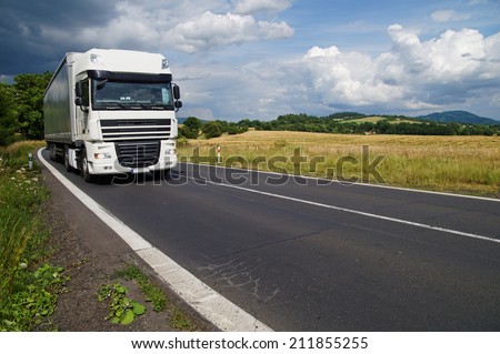 White truck on the road in a rural landscape, in the background of a green cornfield and wooded mountains
