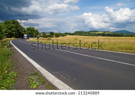 Road in a rural landscape with dramatic clouds in the sky, two trucks leave from the trees bend, in the background cornfield and wooded mountains