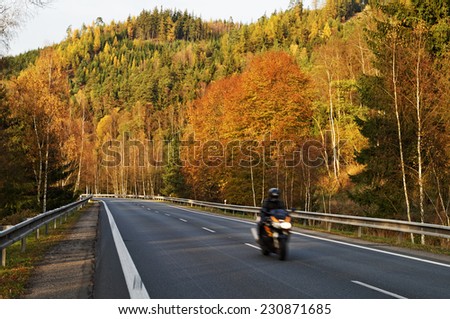 Asphalt road in the autumn landscape with a ride motorcycle, over the road forested mountain, deciduous trees with leaves in bright autumn colors