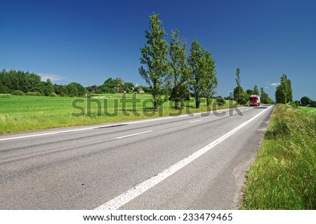 Empty road lined with poplar alley in the countryside, in the distance passing red truck, in the background of green fields, villages, mountain with the ruins of the castle