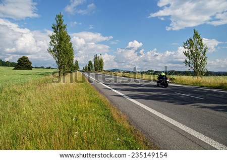 An empty road lined with poplar alley in the countryside, passing motorcycle, in the background green field, forest and mountain