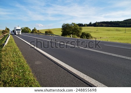 Asphalt road in a rural landscape. The arriving white truck on the road. Meadow and forest in the background.