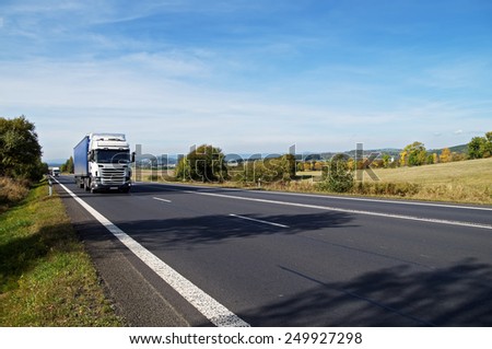 Asphalt road in a rural landscape. The arriving two white trucks on the road. The trees cast shadows on the road.