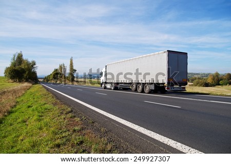 White truck travels on the asphalt road in the countryside. Fields, meadows and trees in early autumn colors in the background.