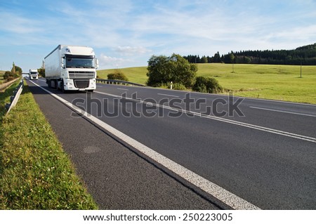 Asphalt road in a rural landscape. The arriving two white trucks on the road. Meadow and forest in the background.