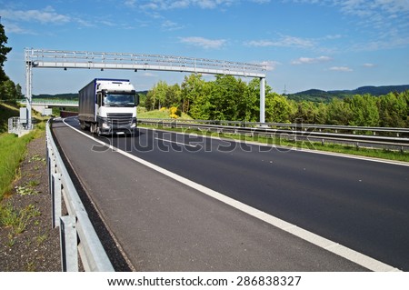 White truck passing through the electronic toll gates on the highway in a wooded landscape. Bridge and forested mountains in the background. White clouds in the blue sky.