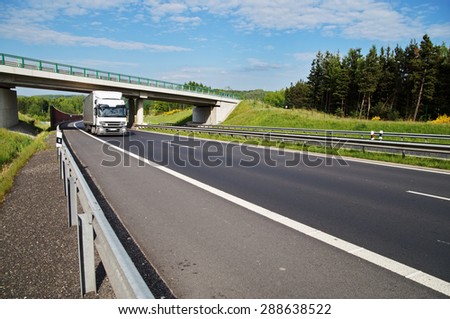 The white truck rides under a concrete bridge over a highway in a wooded landscape. Noise protection wall. White clouds in the blue sky.