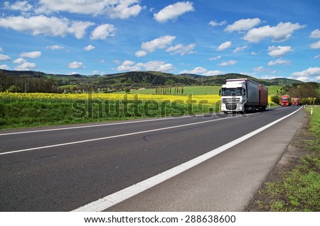 Trucks driving on the asphalt road around the yellow flowering rapeseed field in rural landscape. Wooded mountains in the background. Blue sky with white clouds.