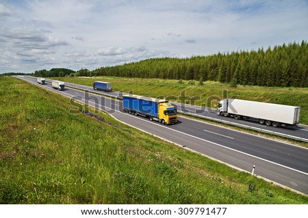 Trucks traveling on an asphalt highway between flower meadows in the countryside. Larch forest on the horizon. Summer sunny day.