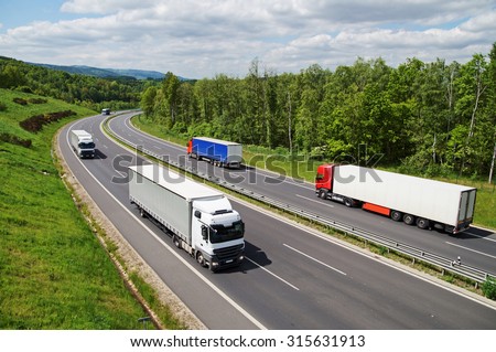 Trucks traveling on an asphalt highway between forests. Wooded mountains in the background. View from above. Sunny summer day.