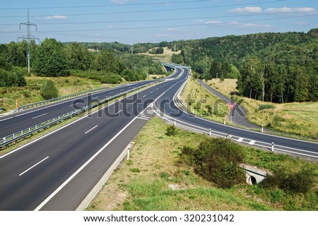 Empty asphalt highway with the slip road and traffic sign give way. Wooded landscape. Drain channel beneath the road. Electronic toll gate in the distance. View from above. Sunny day with blue skies.