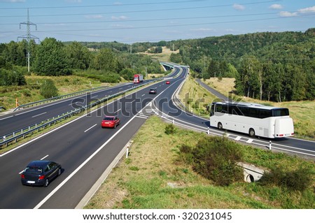 White Bus arriving to the asphalt highway on the slip road in a wooded landscape. Red passenger cars and truck driving on the highway. Electronic toll gate in the distance. View from above.