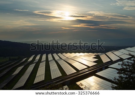 Sunset over the solar power plant between forests. The smoking smokestack and forested mountains fading into the misty haze and smog in the background.