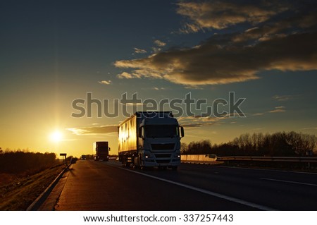 Trucks on asphalt highway in a rural landscape at sunset.