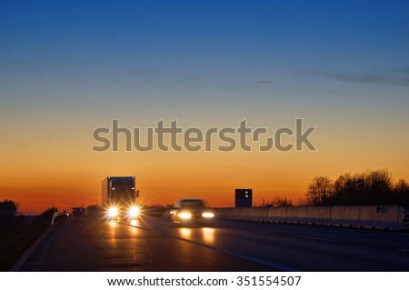 Highway with oncoming trucks and a car after sunset. Shining the spotlight cars. Information boards about the temperature. Blue and orange sky is clear.