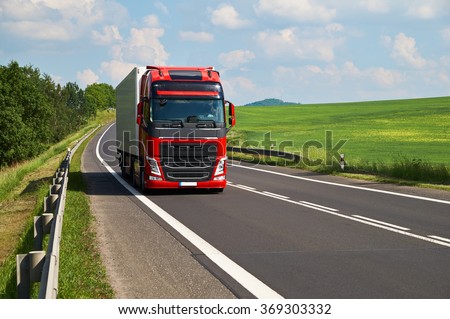 Red truck driving on asphalt road in a rural landscape. Green fields and alleys leading along the road. Sunny summer day with a cloudy sky.