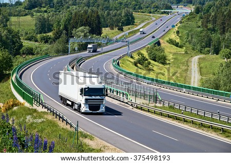 White trucks traveling on the asphalt highway with electronic toll gates in a wooded landscape. View from above. Sunny summer day.