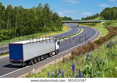 Truck driving on the highway with a double bend in a landscape. The bridge over the highway. Deciduous forest along the highway. View from above. Sunny summer day with blue skies and white clouds.