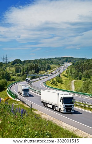 White trucks drive along an asphalt highway with electronic toll gate in a wooded landscape. View from above. Sunny summer day with blue skies and white clouds.