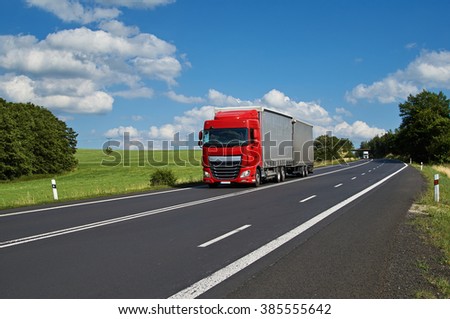 Red truck driving on asphalt road in a rural landscape. White truck coming from afar. Sunny summer day with blue skies and white clouds.