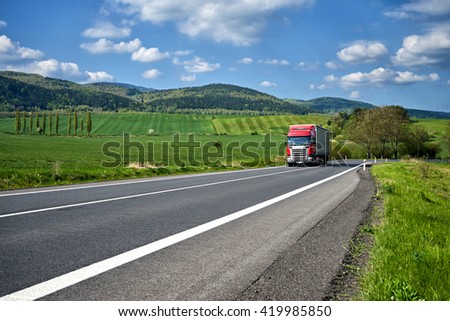 Rural landscape with asphalt road and oncoming red truck. Green fields and wooded mountains in the background. Sunny spring day with blue skies and white clouds.