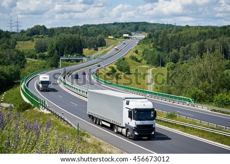 Trucks and cars driving on a highway through a forested hill and a bridge across the valley. Electronic toll collection in the middle of the road. Bridges in the distance. View from above.