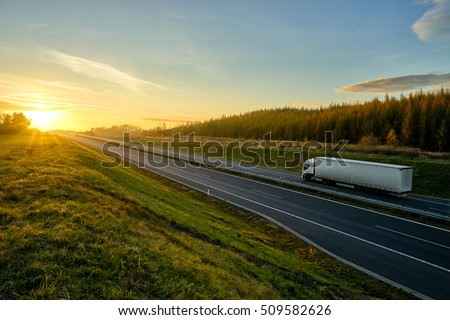 Asphalt highway with a ride white truck between green meadows and larch forest in autumn landscape at sunset.