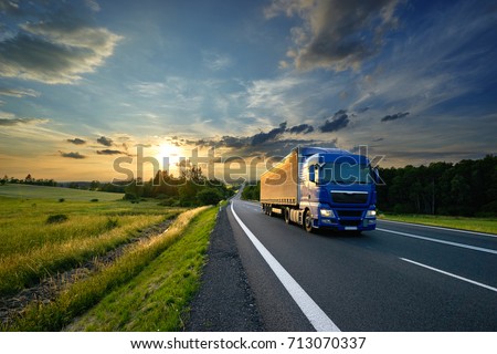Blue truck driving on the asphalt road in rural landscape at sunset