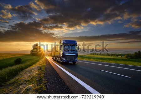 Blue truck driving on the asphalt road in rural landscape at sunset between dark clouds