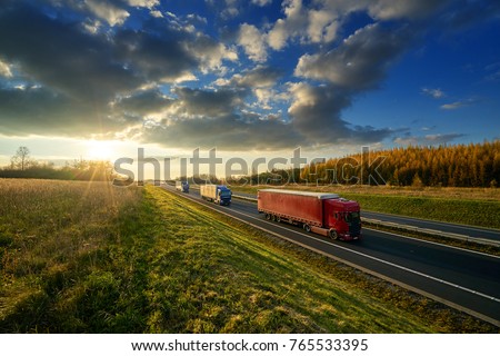 Three trucks driving on a highway in autumn landscape at sunset with dramatic clouds