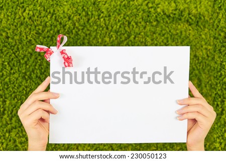 Close-up of a white sheet of paper holding by two woman hands with a red bow,ribbon in a corner isolated on green background.