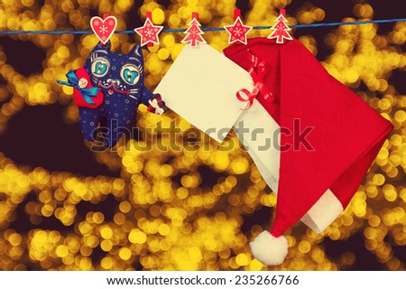 Christmas decoration santa hat and a cat toy and empty sheet of paper with a red ribbon hanging on rope over golden blurred lights background