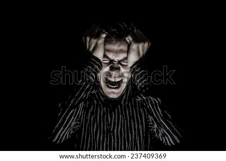 Stressed, aggressive, frustrated portrait of a young student, man,screaming holding his hands up on head isolated on black background.Facial expression