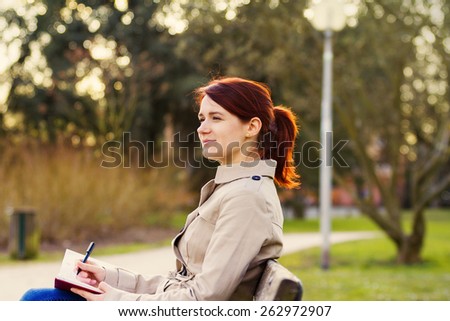 Young smiling businesswoman sitting in a classic city park,student professional outdoors  holding a red ,journal writing in it.Businesswoman smiling,Life style,