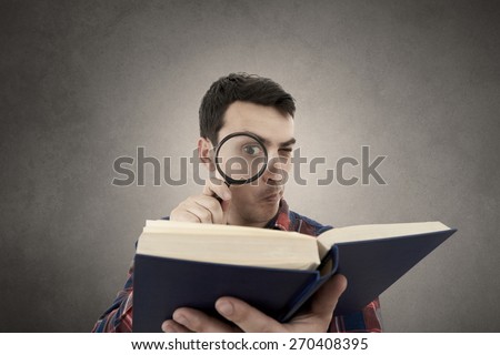 Perplexed Young man student holding magnifying glass and a book isolated over grey background.Curious young student man holding book with a magnifying glass.