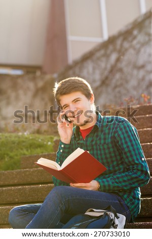 Smiling young man holding book and speaking on mobile phone in a city on stair .Young smiling student  outdoors  with tablet and mobile phone.Life style.City